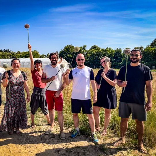 Un groupe de 6 salariés est debout dans un champs. Ils et elles tiennent une fleur dans les mains. Une serre apparaît dans le fond.