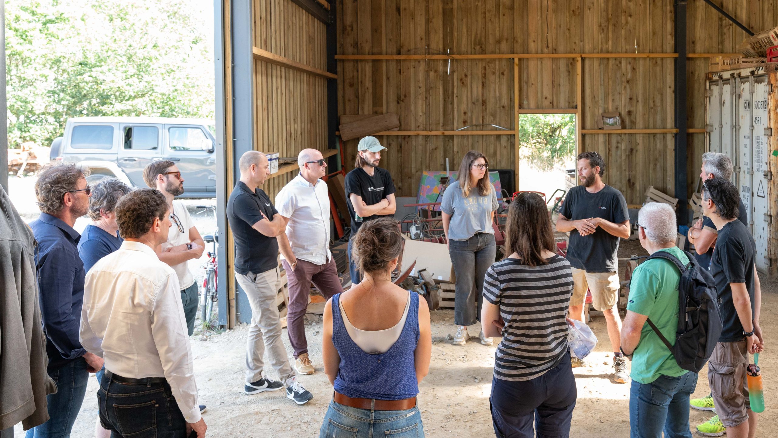 Un groupe de 14 personnes sont debout, en cercle dans un hangar en bois.