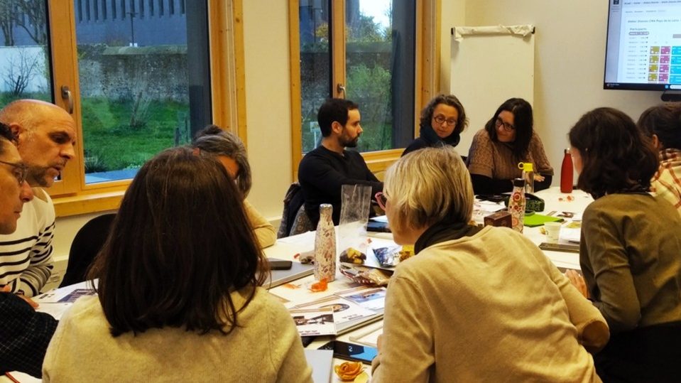 Un groupe de onze salariés est assis autour d'un table. Un écran affiche des diagramme. Les personnes discutent entre eux et regardent des cartes. Ils sont dans une salle de réunion.